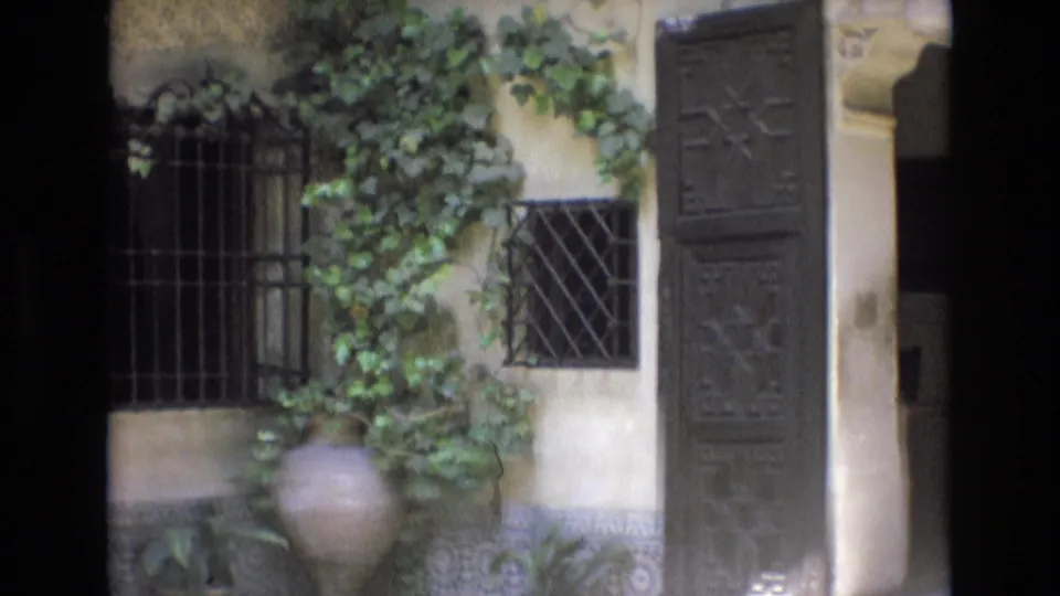 1972 Spain 8mm frame showing a stucco courtyard wall with iron window grilles, ivy, a large terracotta pot, and decorative tilework along the base.