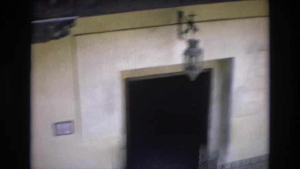 1972 Spain 8mm frame showing a dark recessed doorway beneath a hanging lantern on a pale stucco wall, with a small wall plaque visible nearby.