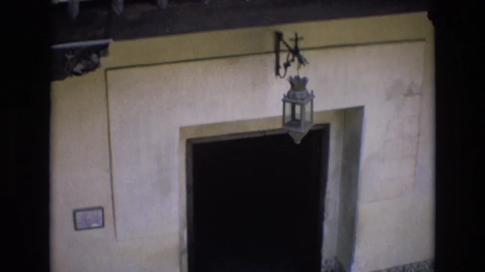 1972 Spain 8mm frame with a closer view of a recessed dark doorway, suspended lantern, pale wall surface, and small rectangular plaque.