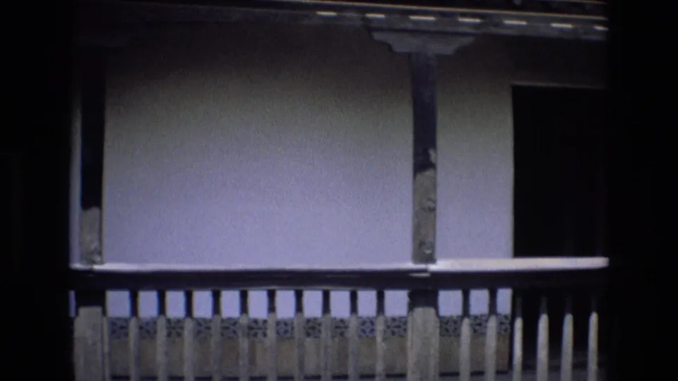 1972 Spain 8mm frame showing a shaded balcony or upper gallery with a decorative railing, vertical supports, pale wall, and dark doorway opening.
