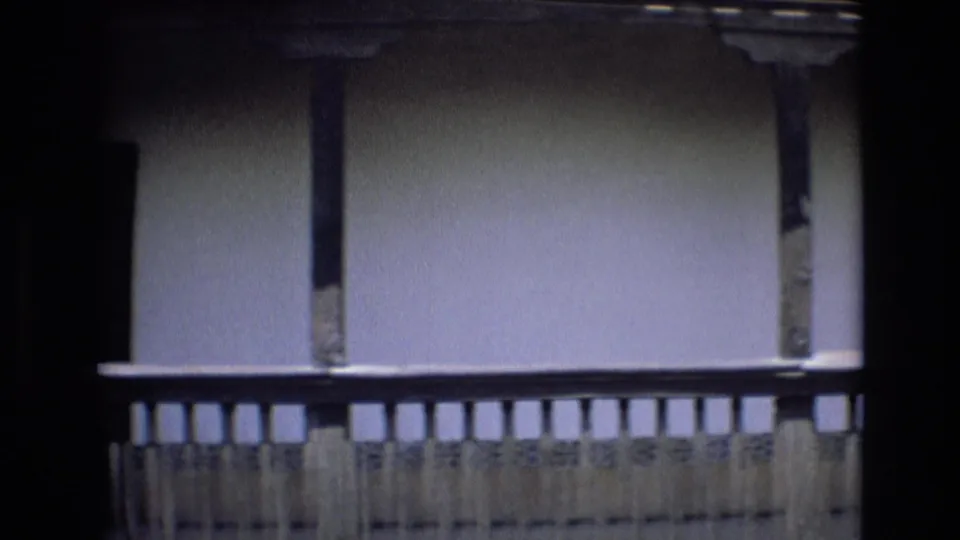 Close 8mm view of a traditional Spanish courtyard balcony with wooden railings, plaster wall, and dark structural shadows.