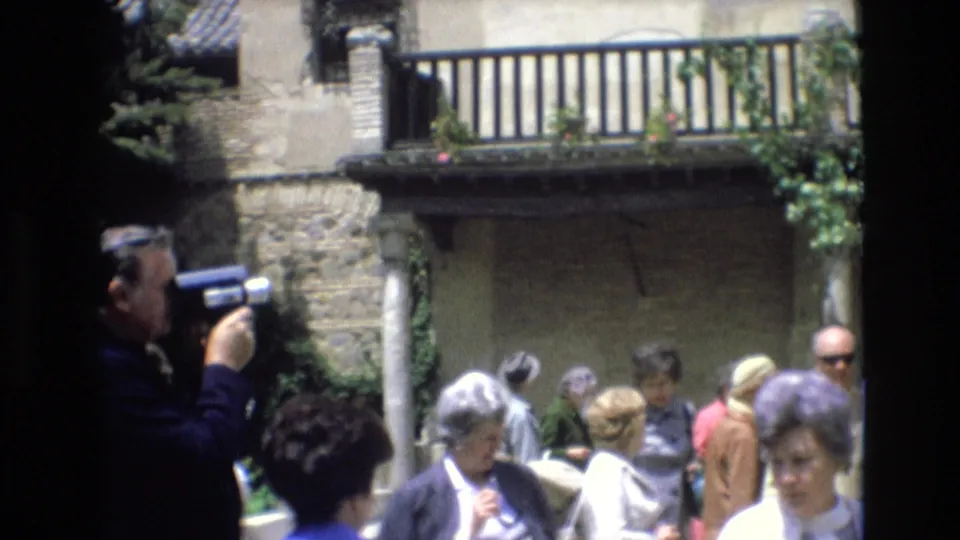 1972 Spain home-movie frame showing tourists in a sunlit courtyard below a wooden balcony, including a man filming at the left edge.