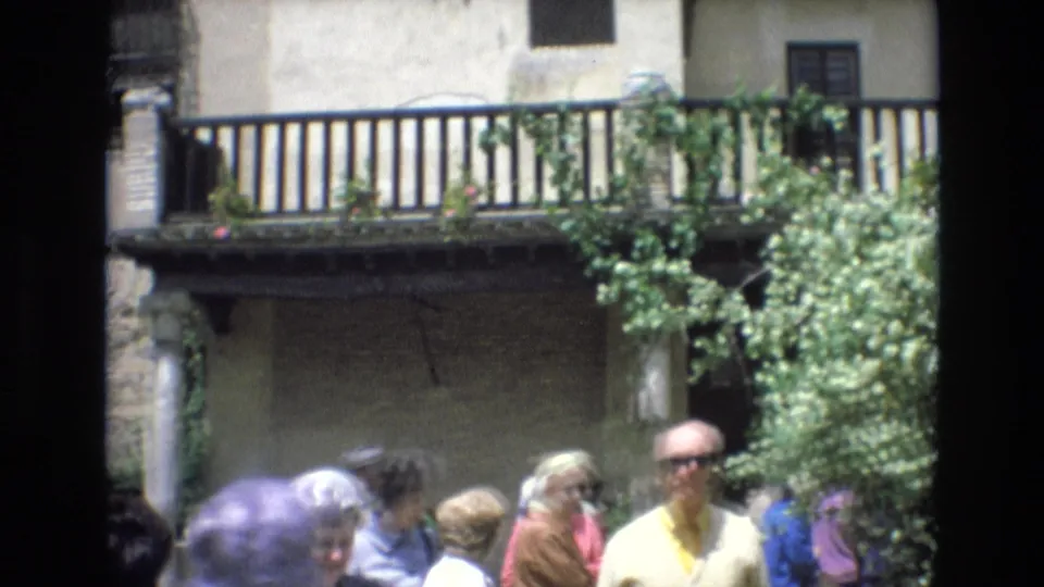 1972 Spain tourist courtyard scene with visitors moving below a wooden balcony, stonework, pale plaster, and climbing greenery.