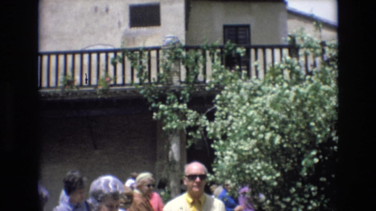 In 1972 Spain, adult tourists move through a sunlit courtyard with dense foliage, a stucco building, and a wooden balcony in the background.