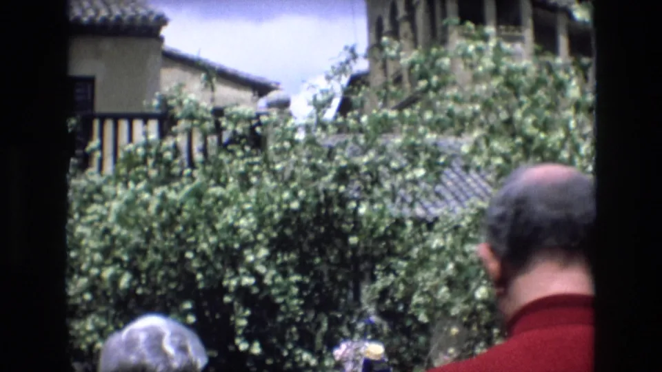 A handheld 8mm frame from Spain shows a visitor in a red shirt from behind, with dense foliage, balcony rails, and tiled roof details beyond.