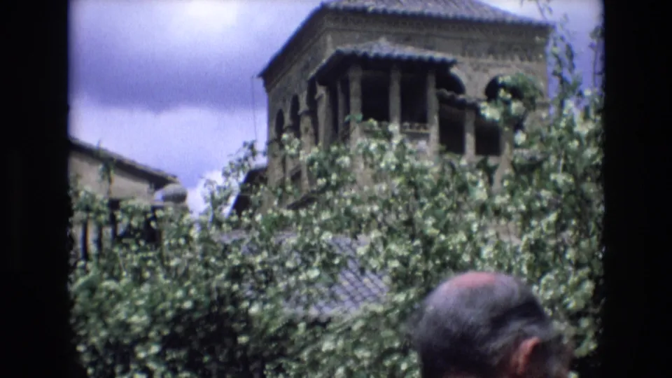 A Moorish-style tower and tiled roof appear above thick courtyard foliage while adult visitors move through the foreground in 1972 Spain.