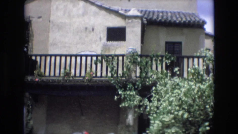 A sunlit Spanish village building with clay roof tiles, a dark window, balcony railing, and greenery appears in the 1972 8mm travel footage.