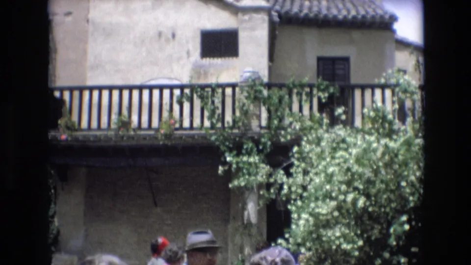 The frame shows a Spanish village building with a balcony and tiled roof above a partly visible crowd of tourists in hats at the lower edge.