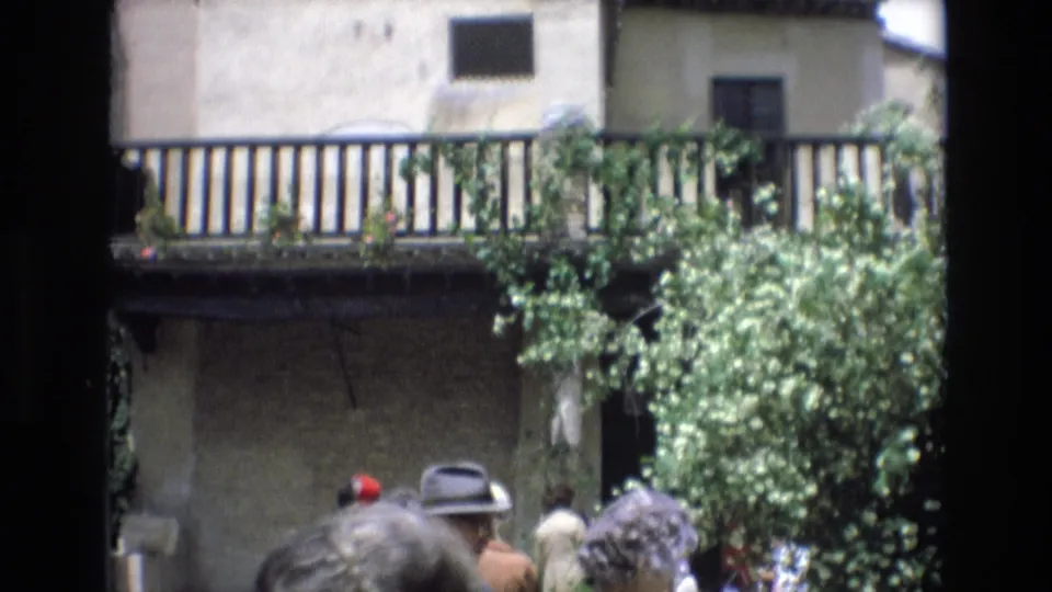 Tourists gather in the lower foreground of this Spain 1972 8mm frame, with brimmed hats visible beneath a vine-covered balcony and sunlit village facade.