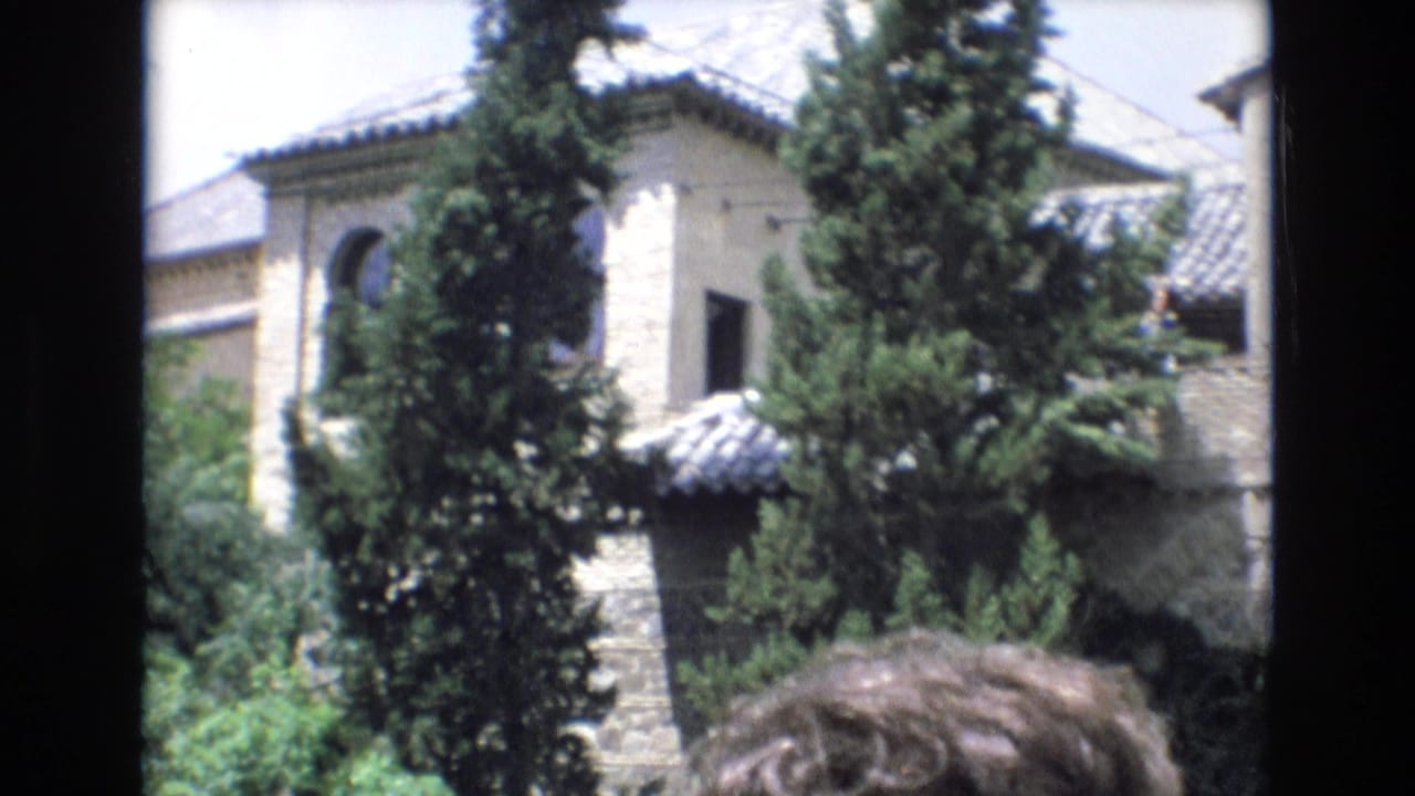 1972 Spain 8mm frame showing a sunlit stone building with tiled roofs, an arched opening, tall conifer trees, and visitors partly visible at the bottom edge.