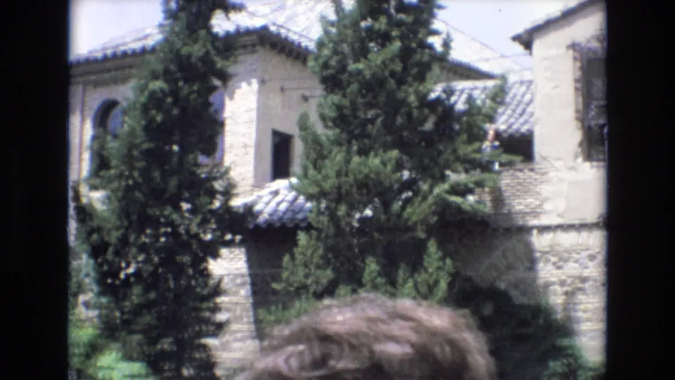 1972 Spain home-movie frame with a pale stone facade, clay-tile rooflines, dark window opening, dense conifer trees, and a cropped visitor in the foreground.