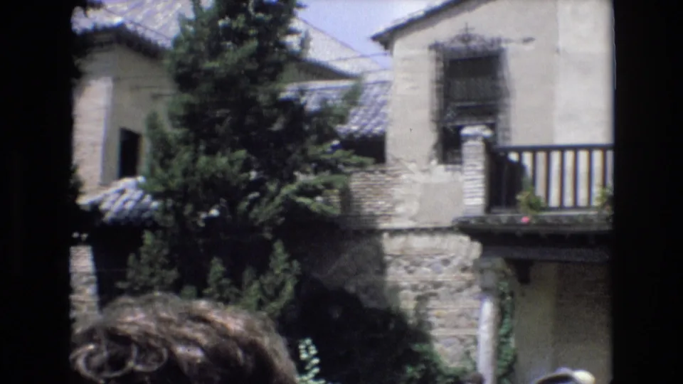 1972 Spain 8mm frame showing a stone and stucco building with a barred window, balcony railing, roof tiles, conifer trees, and cropped visitors near the camera.