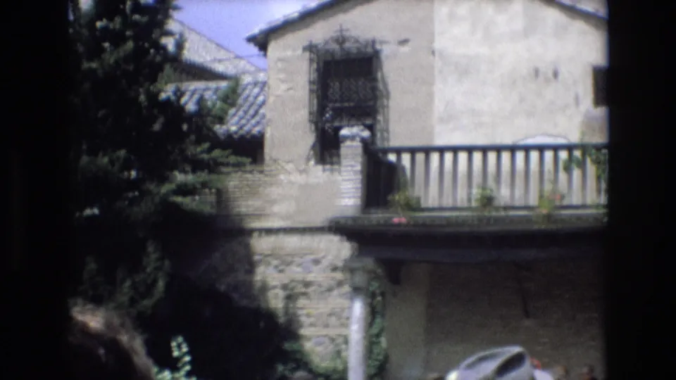 1972 Spain home-movie frame with a black metal balcony railing, barred upper window, stone lower wall, leafy trees, and a cropped foreground visitor.