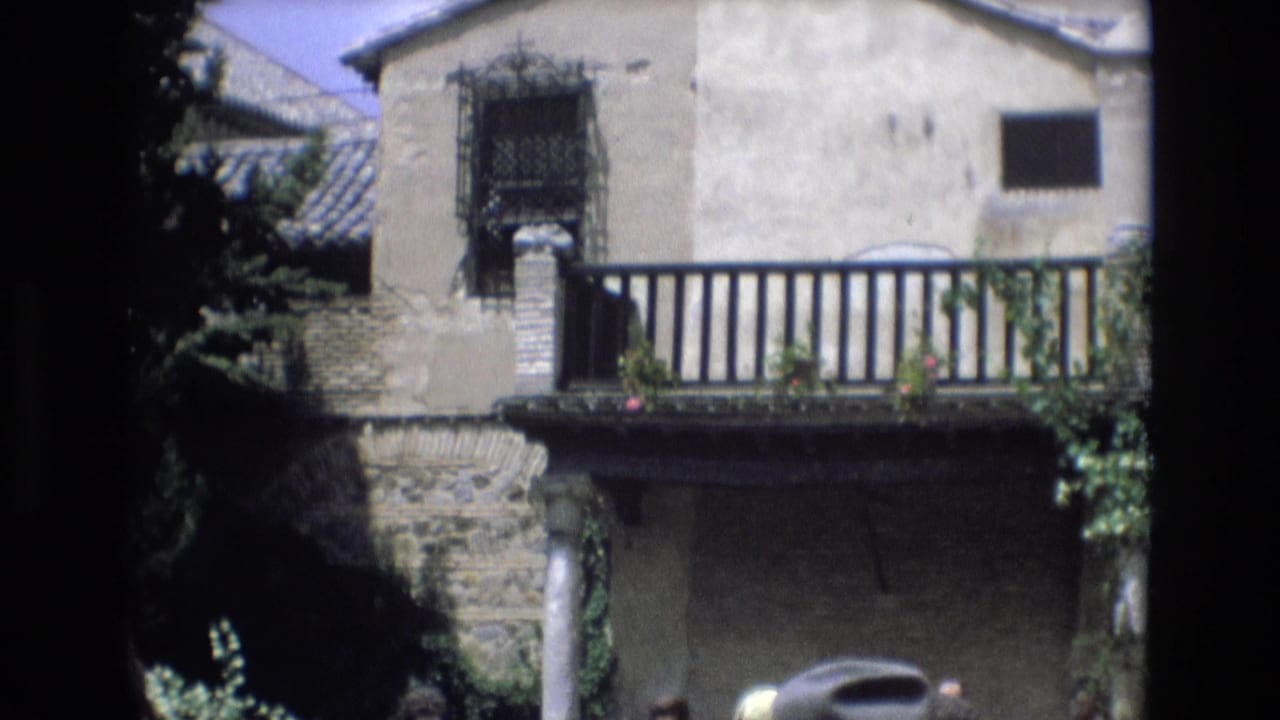 1972 Spain 8mm frame showing a weathered stone and stucco villa facade with black balcony railings, iron window grilles, roof tiles, and visitors partly visible at the bottom edge.
