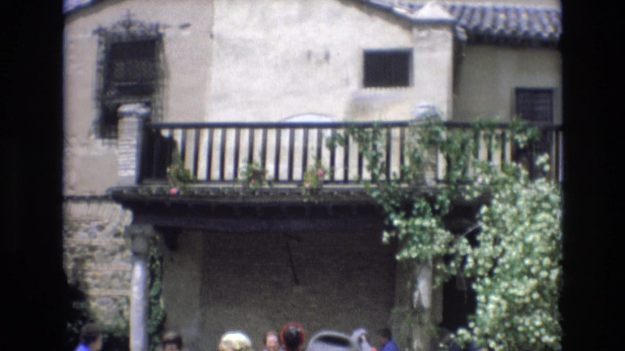 1972 Spain 8mm frame of an old villa exterior with a long balcony, dark window grilles, green vines, and several tourists partly seen below the railing line.