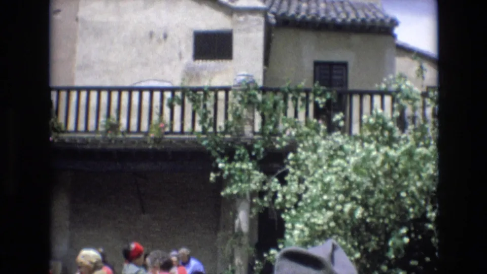 1972 Spain 8mm frame with the camera panned across balcony railings, tiled roof, a leafy shrub in bloom, and tourists partially visible near the shaded courtyard edge.