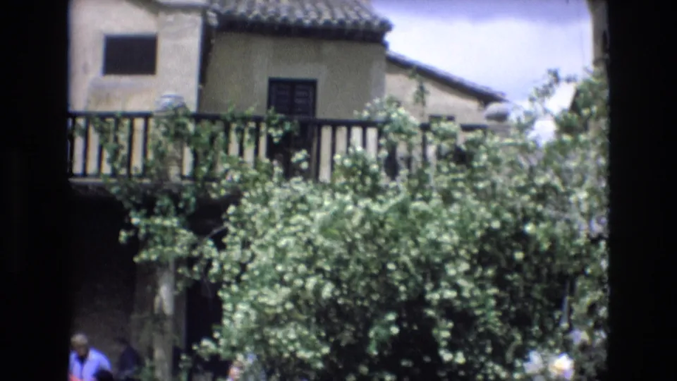 1972 Spain 8mm frame showing dense courtyard greenery in front of a traditional villa balcony, with the tiled roof and partially visible tourists near the lower edge.