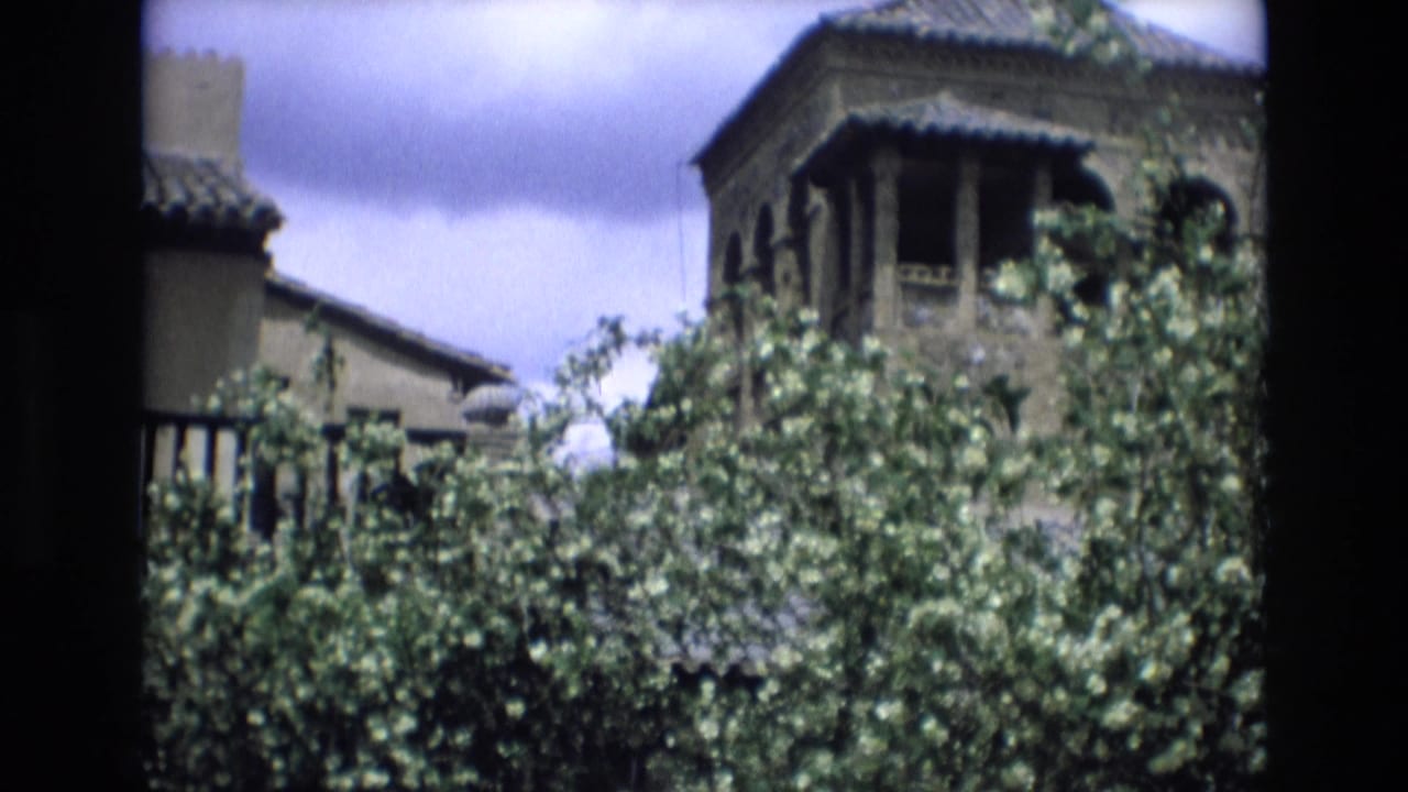 Leafy trees frame historic-looking buildings with tiled roofs and an upper balcony or tower.