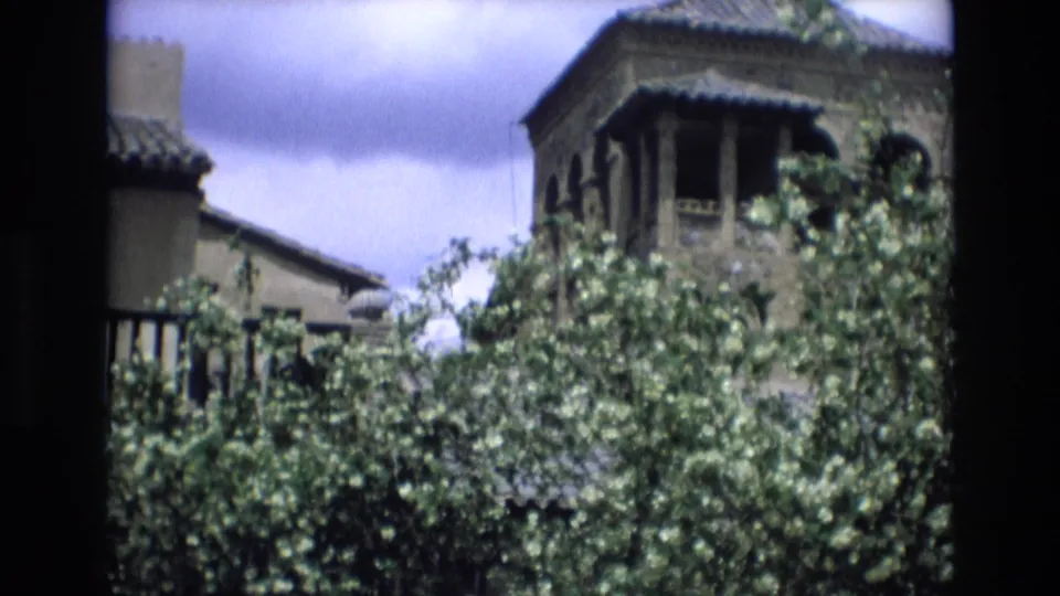 1972 Spain frame showing dense greenery in front of old street architecture with tiled roofs and an elevated balcony or tower detail.