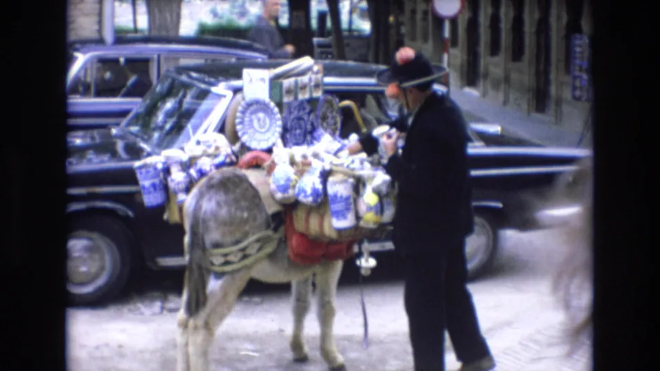 1972 Spain street frame showing a man in a dark suit and hat beside a donkey carrying blue and white ceramics, with dark parked cars and buildings nearby.