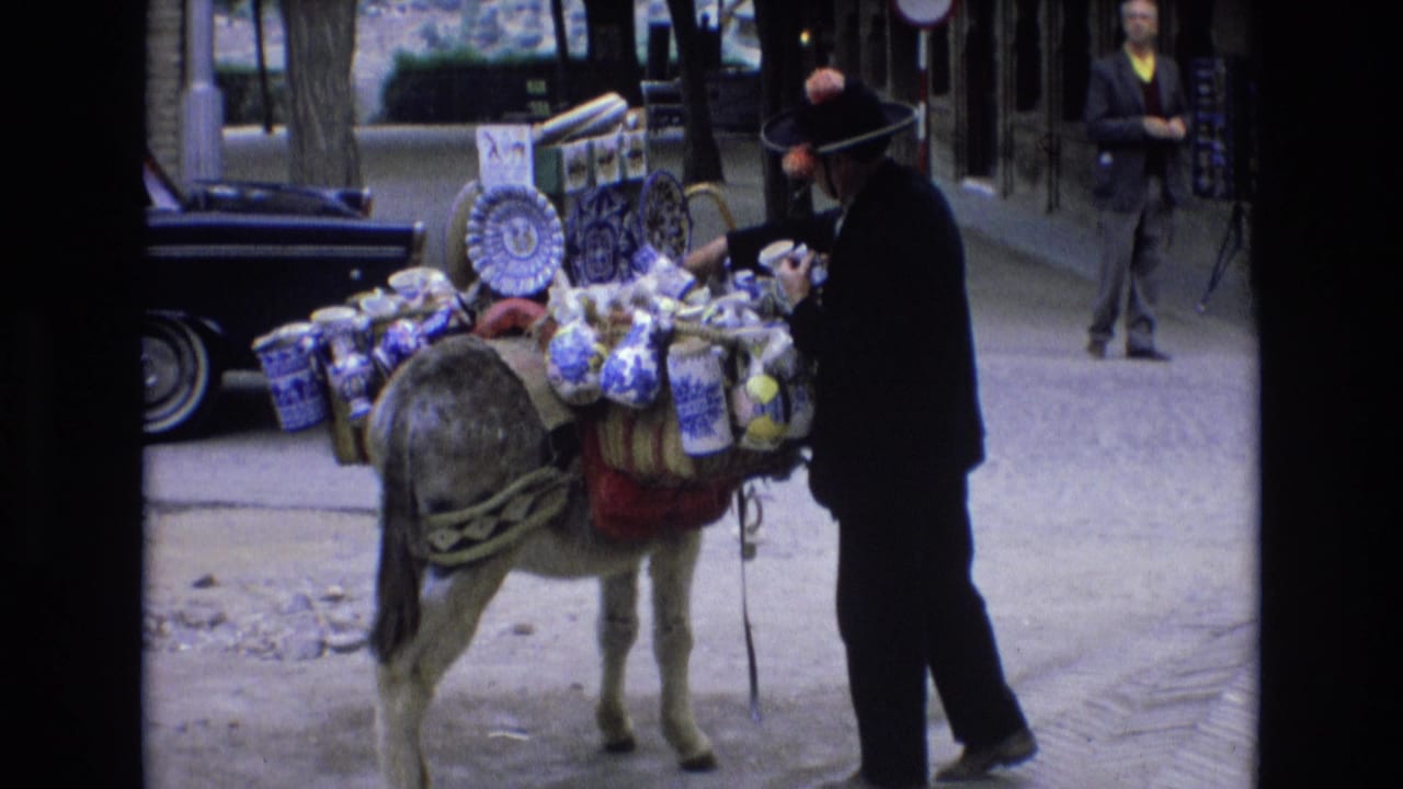 In supplied 1972 Spain footage, a street vendor in a dark suit and brimmed hat stands beside a donkey carrying blue-and-white ceramic goods and bundled merchandise.