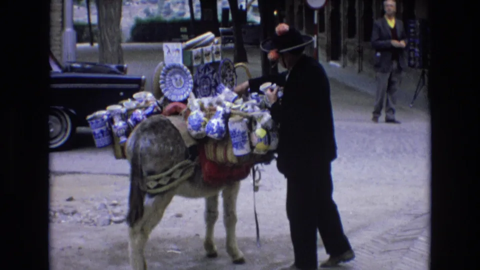 In supplied 1972 Spain footage, a street vendor in a dark suit and brimmed hat stands beside a donkey carrying blue-and-white ceramic goods and bundled merchandise.