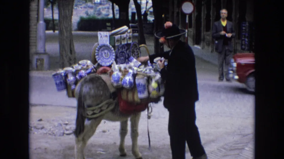 The loaded donkey remains beside the suited vendor while a red car moves through the background of the 1972 Spain street scene.