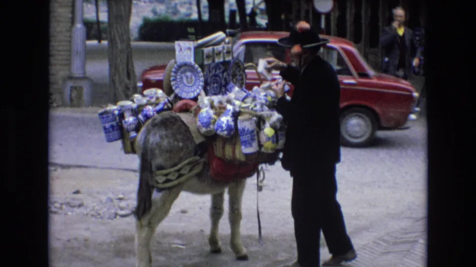 A vendor appears to handle or adjust goods on the donkey while the red car passes behind, with street trees and a pedestrian visible farther back.