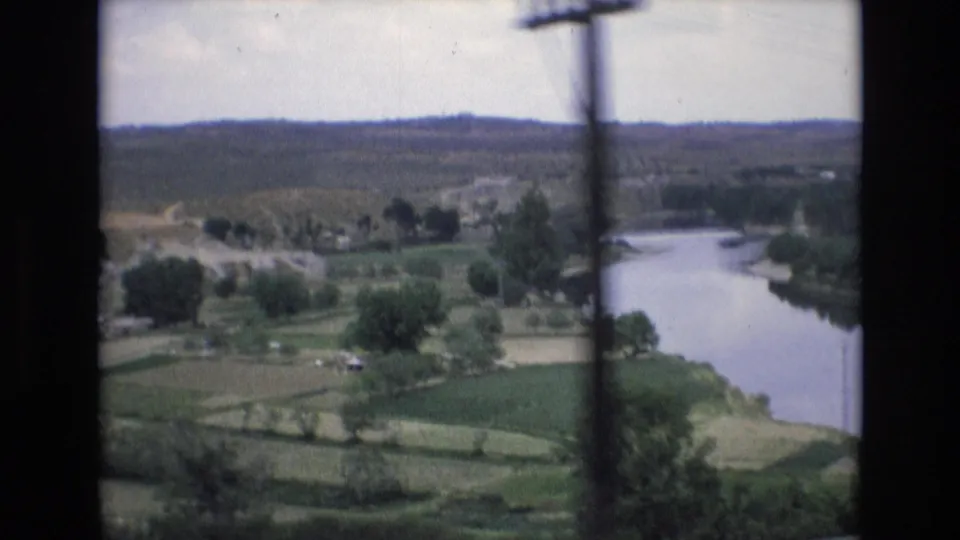 1972 Spain 8mm frame showing a winding river beside green fields, scattered trees, a road, and distant hills from an elevated moving viewpoint.