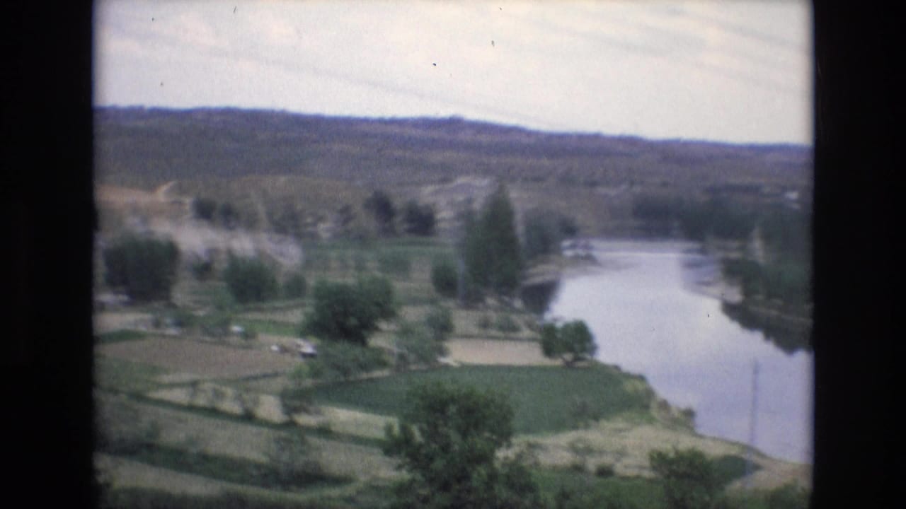 1972 Spain 8mm frame showing a winding river through green fields with scattered trees, distant hills, and faint utility lines in a hazy sky.
