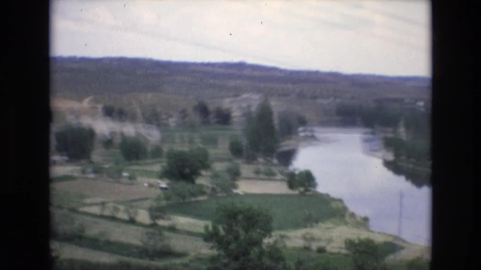 1972 Spain 8mm frame showing a winding river through green fields with scattered trees, distant hills, and faint utility lines in a hazy sky.