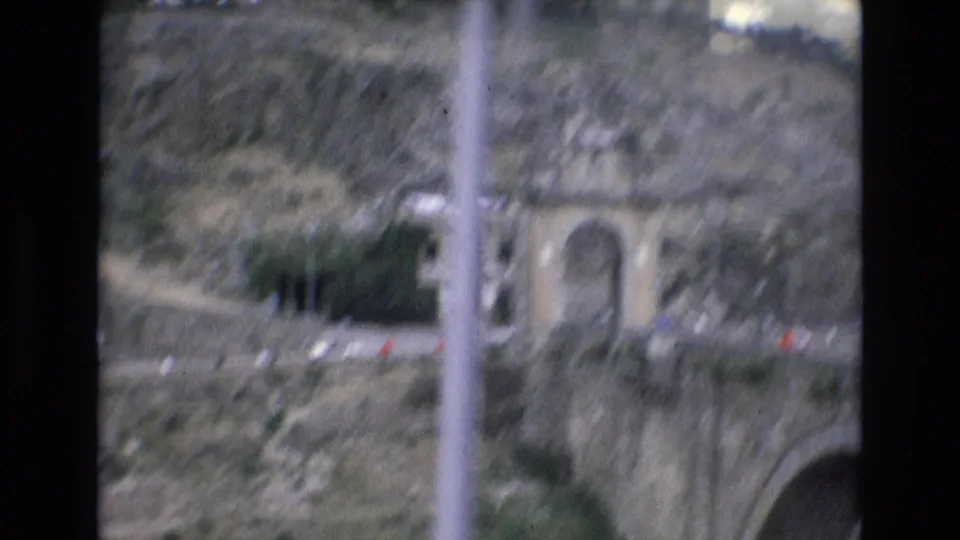 1972 Spain 8mm frame showing a stone archway set into a rocky hillside, with a road or bridge structure below and small distant figures or vehicles near the entrance.