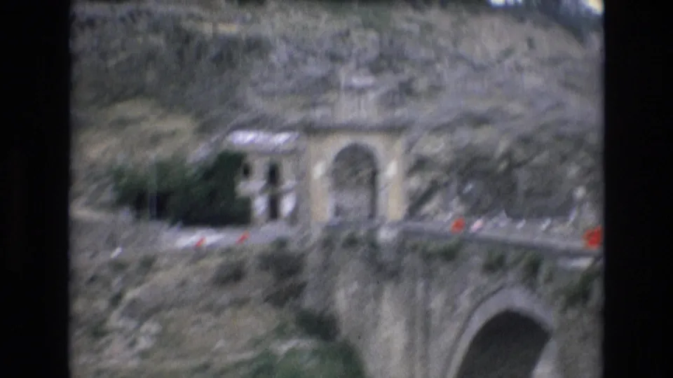 1972 Spain sightseeing frame with a stone archway in a steep rocky slope, small distant people or vehicles along the road, and part of a larger bridge arch below.