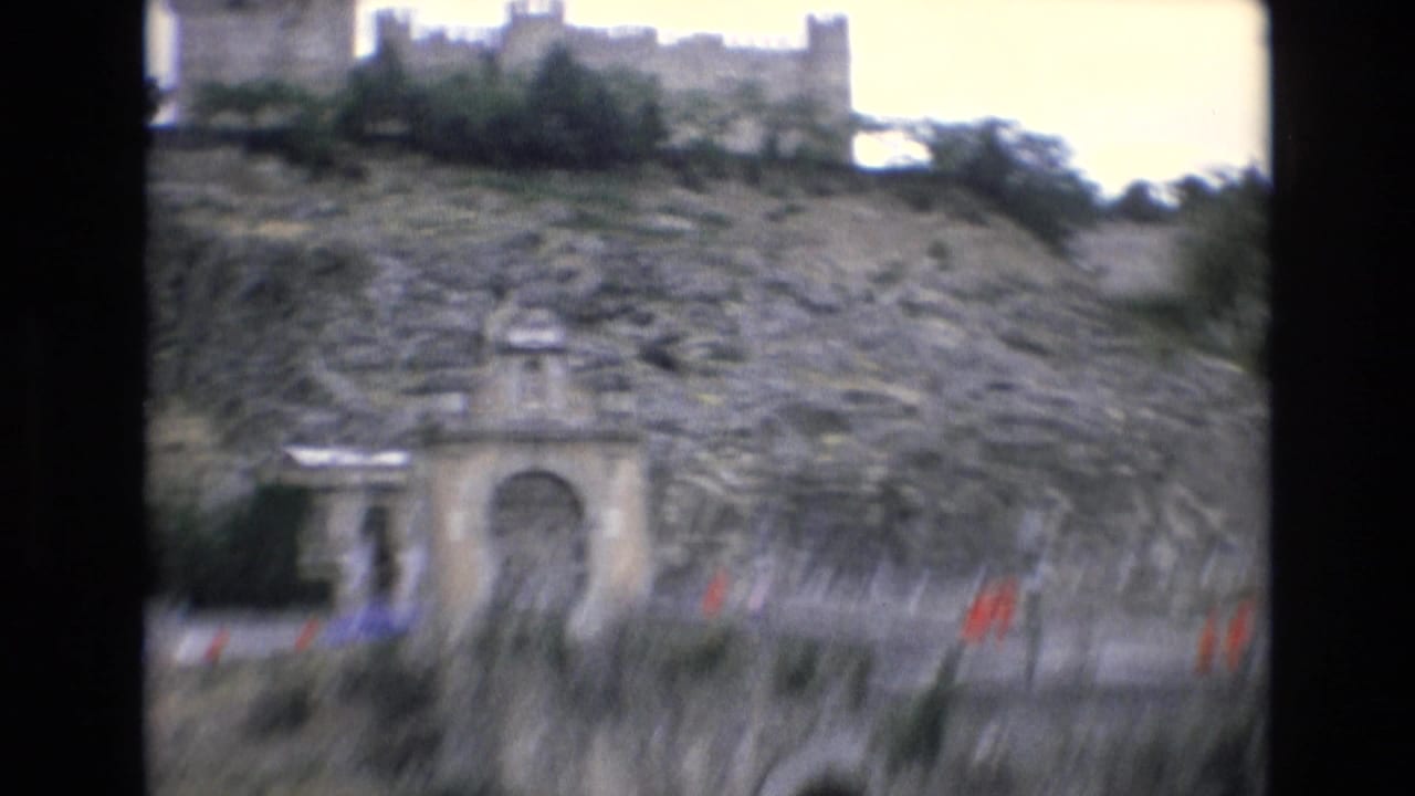 1972 Spain 8mm frame showing a stone bridge and arched gateway set into a rocky hillside, with distant visitors in red shirts and a fortress-like structure above.