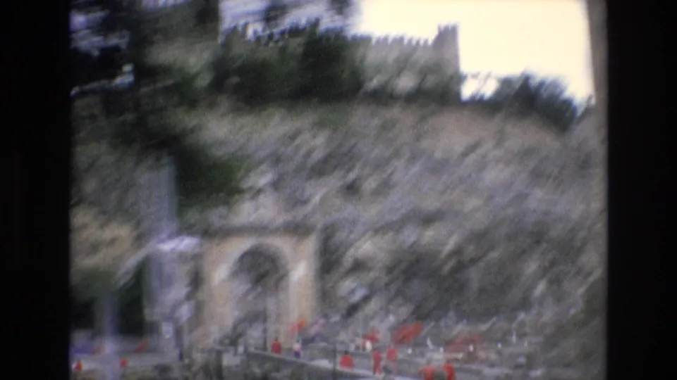 1972 Spain 8mm frame of an arched stone entrance at the base of a rocky cliff, with small groups of people visible near the bridge and ridge structure above.