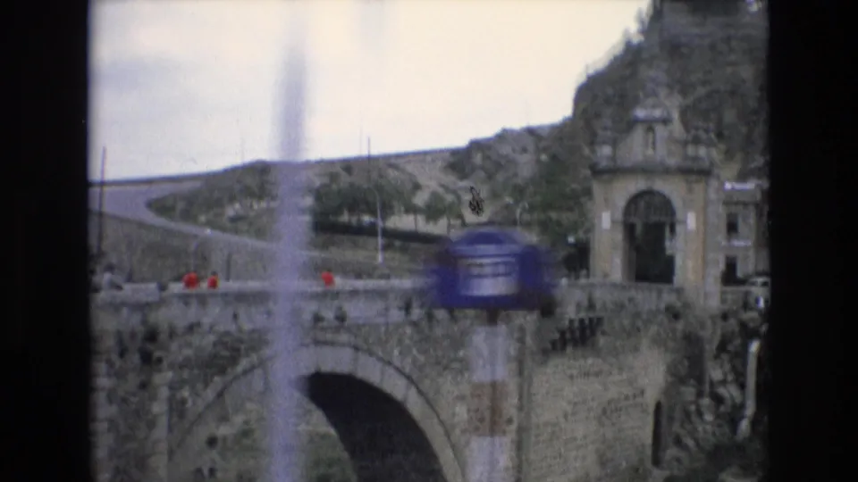 1972 Spain 8mm frame showing a stone bridge and road with a blue car passing near an arched gateway, rocky hillside, and scattered visitors in red shirts.