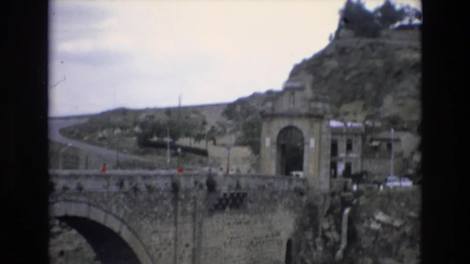 1972 Spain 8mm frame with a stone bridge in the foreground, an arched gateway at right, a road curving up the rocky hillside, and distant visitors visible near the entrance.