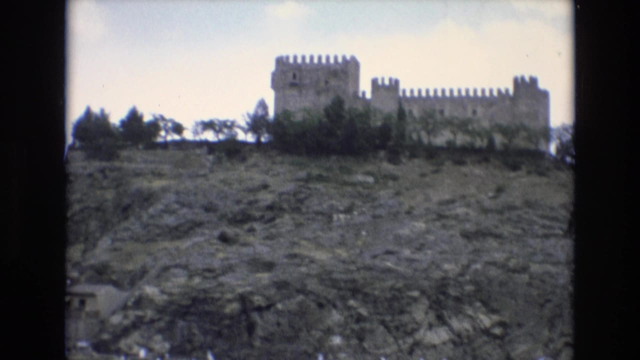 1972 Spain 8mm frame showing a distant crenellated stone castle above a rough rocky slope with trees along the ridge.