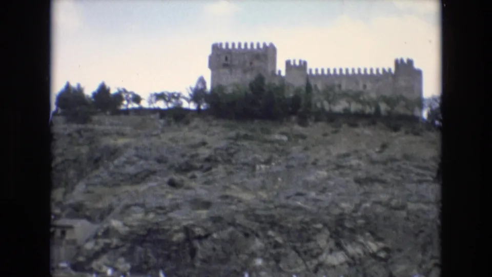 1972 Spain 8mm frame showing a distant crenellated stone castle above a rough rocky slope with trees along the ridge.