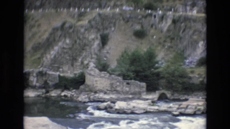 1972 Spain 8mm frame looking toward river rapids, low stone ruins, an arched masonry opening, and rocky terrain rising behind.