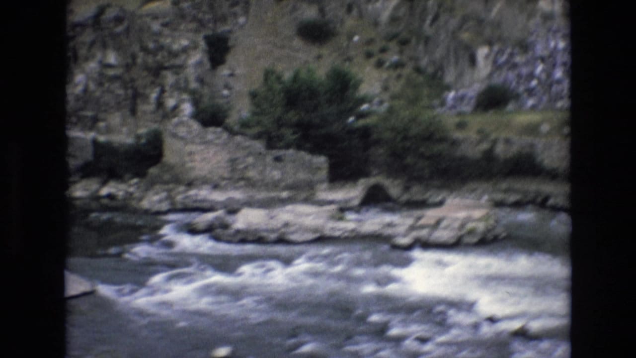 1972 Spain 8mm frame showing a fast-flowing river beneath weathered stone walls, rocks, shrubs, and a hillside bank.