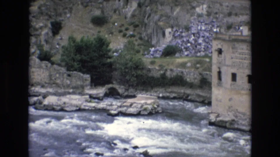 1972 Spain 8mm frame with rushing water, old stone walls, an arched opening near the bank, trees, and a tall masonry building at the right edge.
