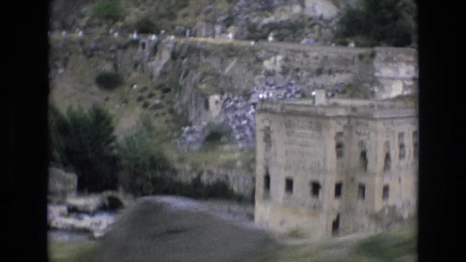 1972 Spain 8mm frame showing a weathered multi-window stone building near the river, with terraced hillside, rubble, shrubs, and old walls behind it.