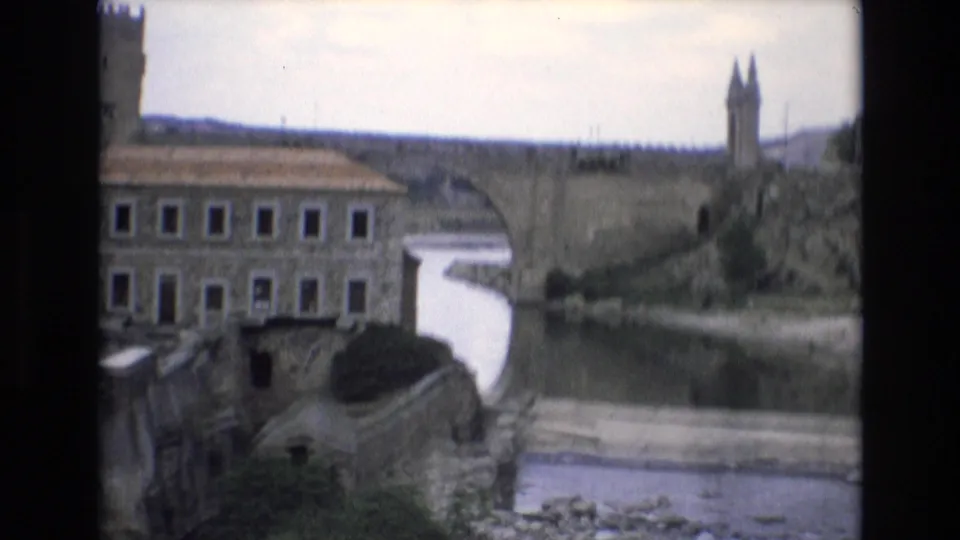 1972 Spain 8mm frame with a stone bridge or passage over calm water, a long masonry building, and a tall fortified-looking tower or wall in the distance.