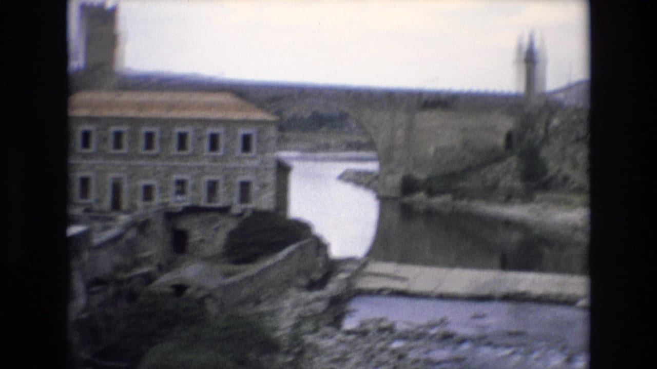 1972 Spain home-movie frame showing a waterside building, stone bridge or river crossing, calm river, and hilltop fortification in soft 8mm color.