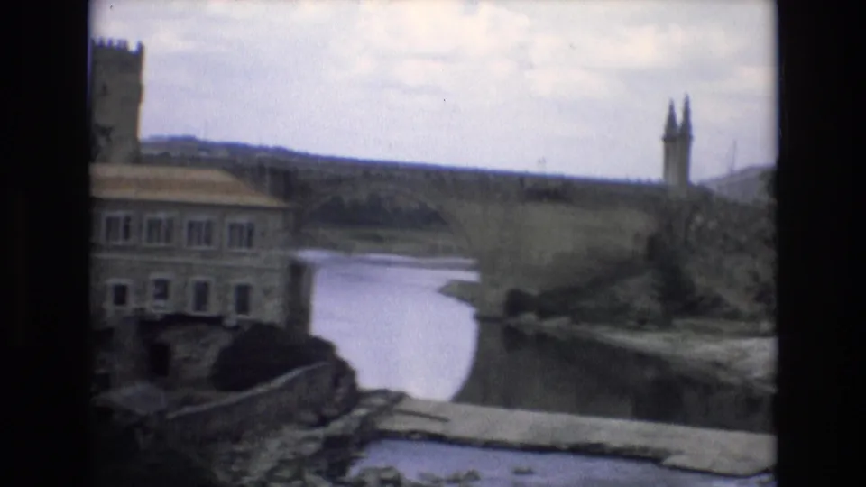1972 Spain travel footage frame with a river bend, waterside building, stone crossing, and fortress-like walls rising behind the town.