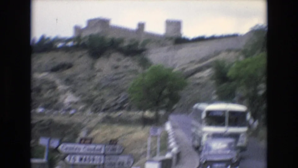 1972 Spain home-movie frame showing a white bus traveling on a winding road beneath rocky hilltop fortifications, with roadside direction signs in the foreground.