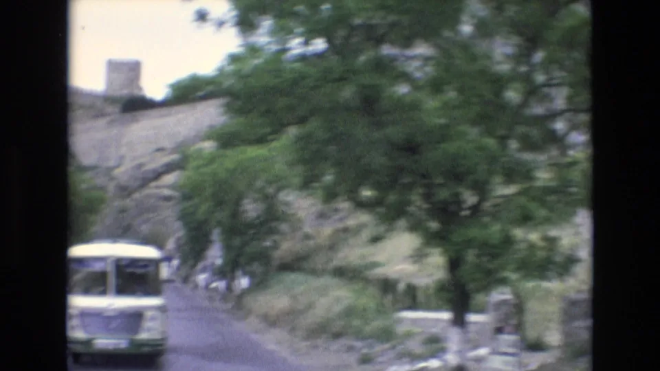 1972 Spain travel frame showing a white bus on a tree-lined road below rocky terrain and a tower-like fortified structure, filmed with soft handheld 8mm texture.
