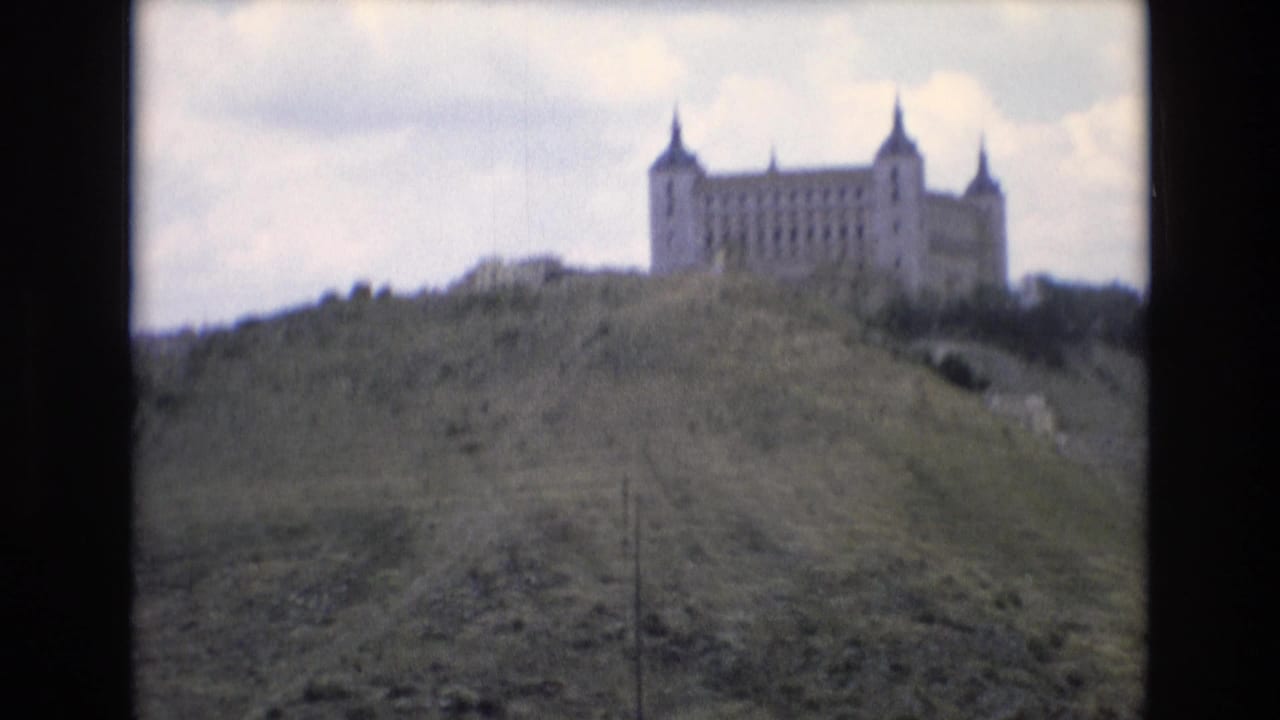 1972 Spain 8mm frame showing a distant castle-like stone building with corner turrets on a grassy hillside under cloud cover.