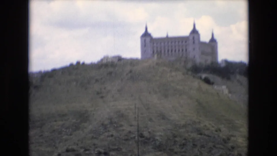 1972 Spain 8mm frame showing a distant castle-like stone building with corner turrets on a grassy hillside under cloud cover.